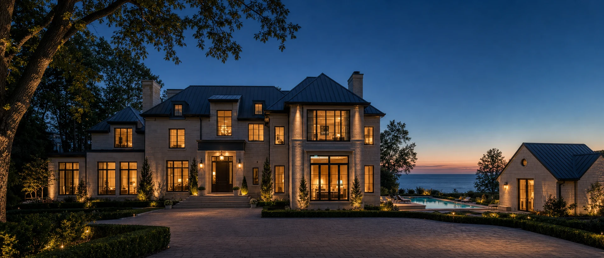 Large Oakville lakeshore estate at twilight with warm interior lighting visible through walls of glass, lit pool deck and pool house at one side of the property, mature trees framing the lot, suggestion of Lake Ontario beyond