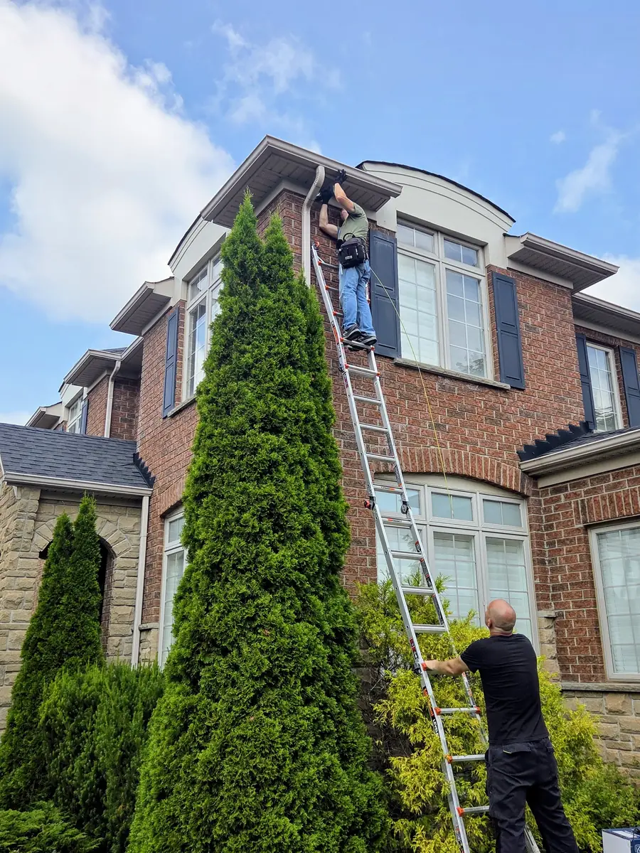 SetupTeam technician on an extension ladder feeding outdoor-rated Cat6 cable up behind the soffit of a two-storey Oakville home while a second technician foots the ladder