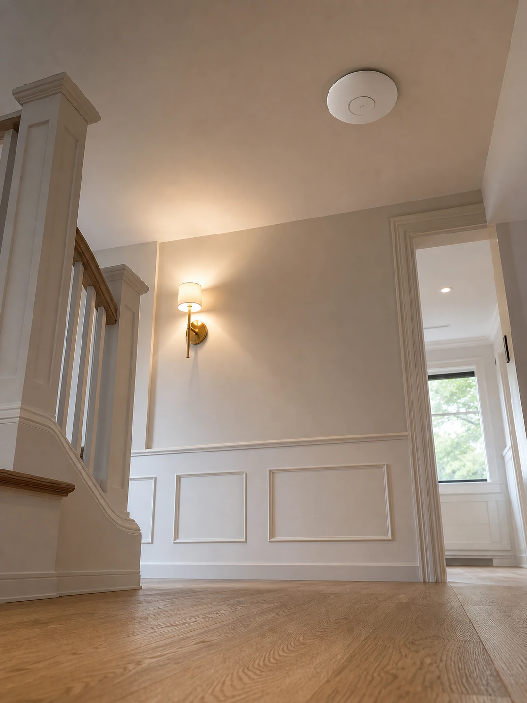 Ceiling-mounted UniFi Wi-Fi access point on the second-floor stair landing of a North York custom infill home with painted millwork walls, hardwood floor, and warm sconce lighting