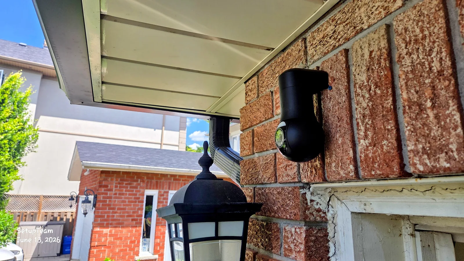 Black UniFi dome-style security camera mounted under a soffit on a residential brick wall, overlooking neighbouring homes