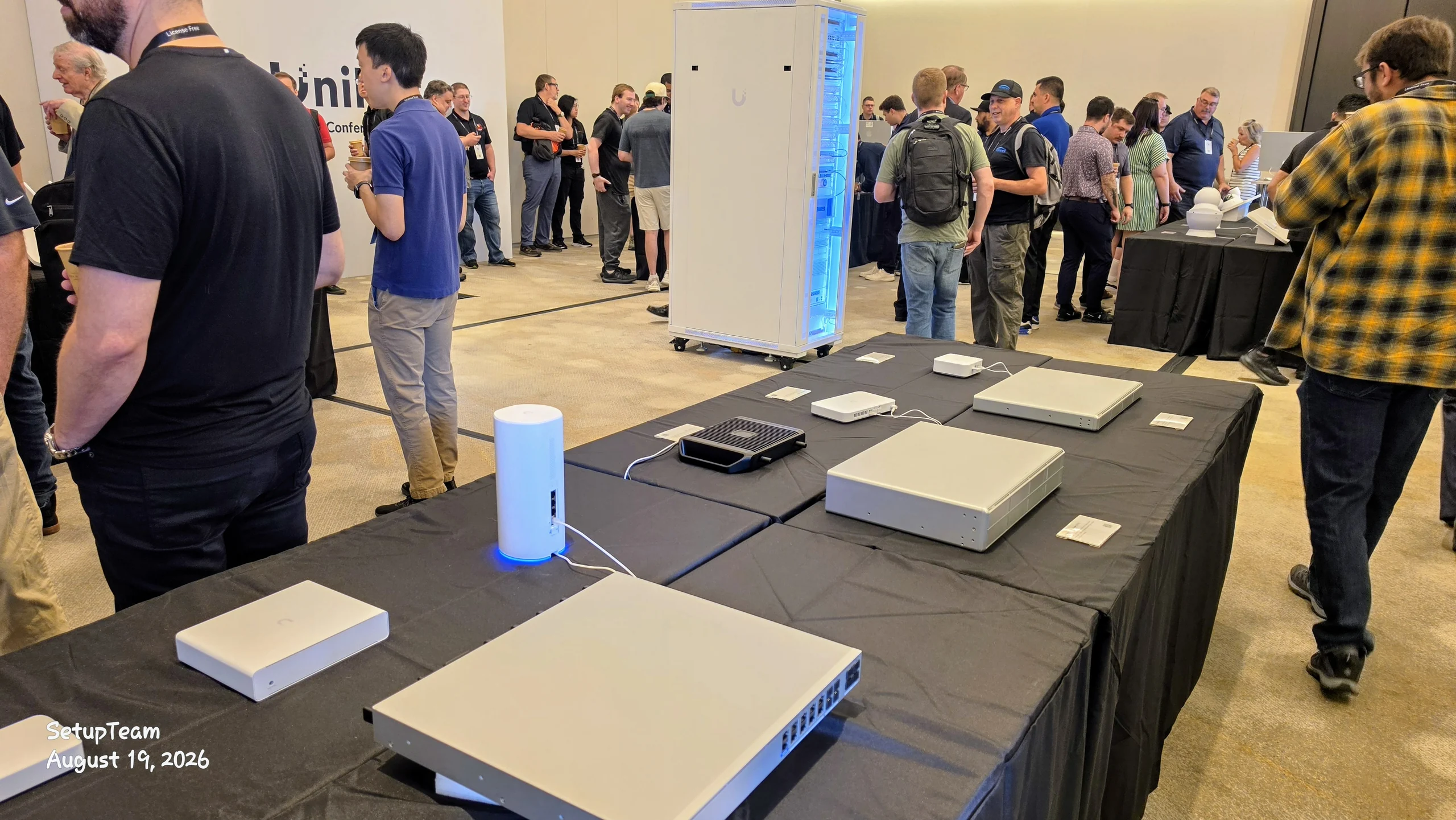 UniFi gateways, switches and access points laid out on black-draped tables for hands-on inspection, with a free-standing lit equipment rack behind