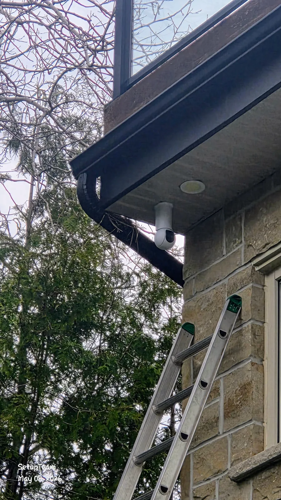 White UniFi cylindrical security camera mounted under the soffit of a stone home, with a clean concealed cable entry